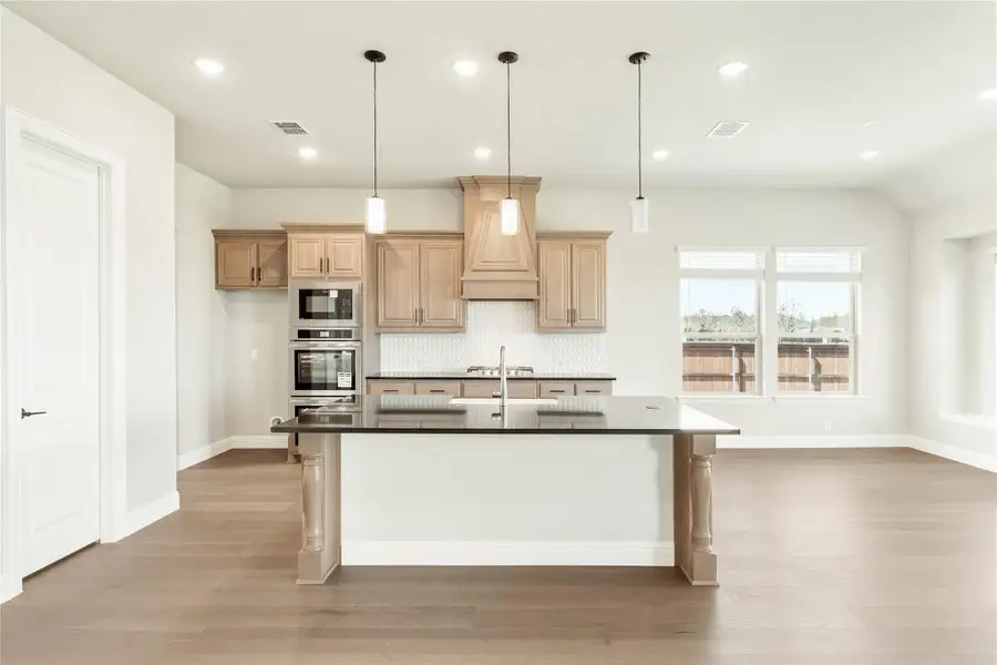 Kitchen with large island, dark countertops, light wood cabinets, pendant lights, and stainless steel appliances Kitchen with large island, dark countertops, light wood cabinets, pendant lights, and stainless steel appliances