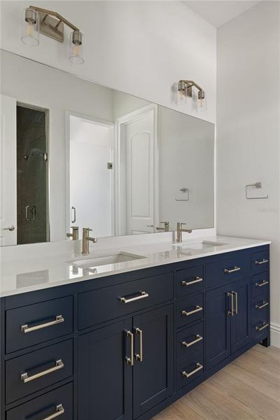 Downstairs primary bathroom vanity featuring dual sinks, a dark blue cabinet with ample storage, a white countertop, modern hardware, and a large mirror reflecting the bright and spacious en suite.