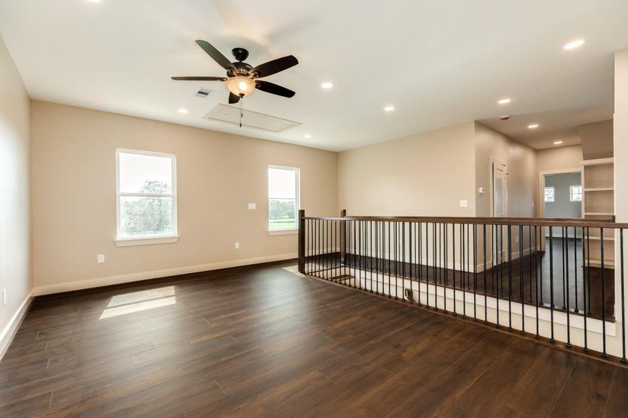This spacious, open area features rich wood flooring, neutral walls, and ample natural light. It includes a ceiling fan, recessed lighting, and a modern railing overlooking a lower level, creating an inviting and versatile space.