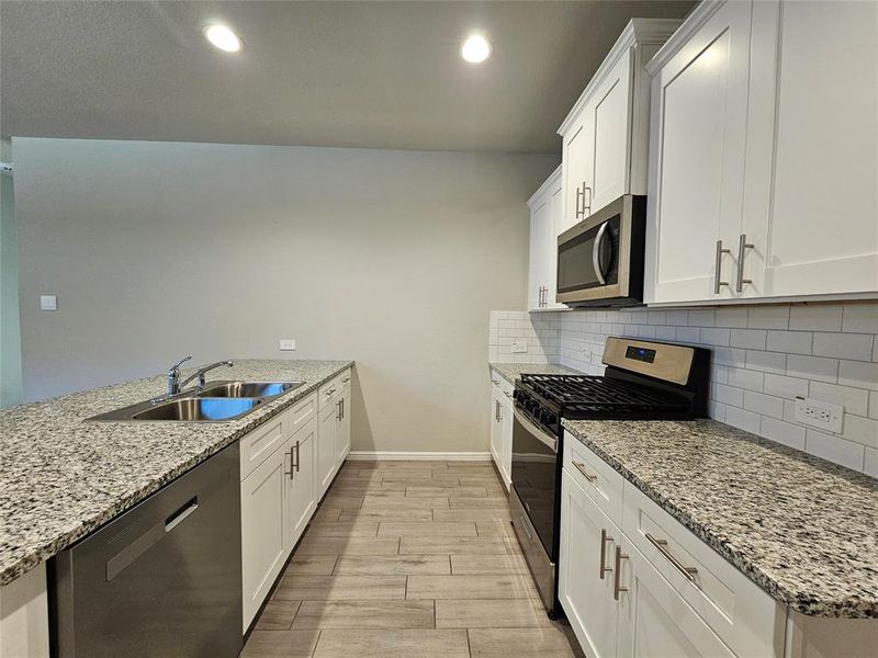 Kitchen with white cabinetry, open floor plan, stainless steel appliances, recess lights, granite counters, backsplash and wood looking tiled floors.