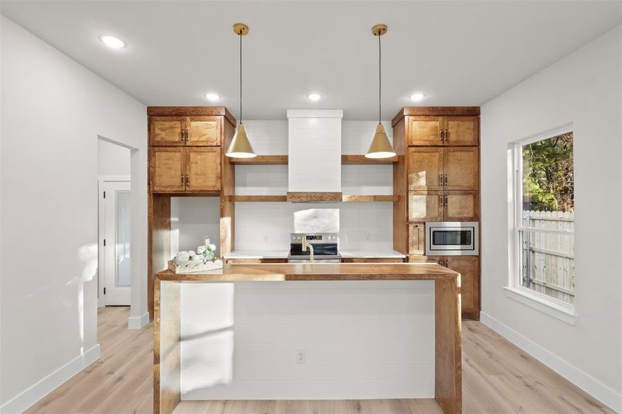 Kitchen featuring pendant lighting, exhaust hood, light wood-type flooring, an island with sink, and appliances with stainless steel finishes