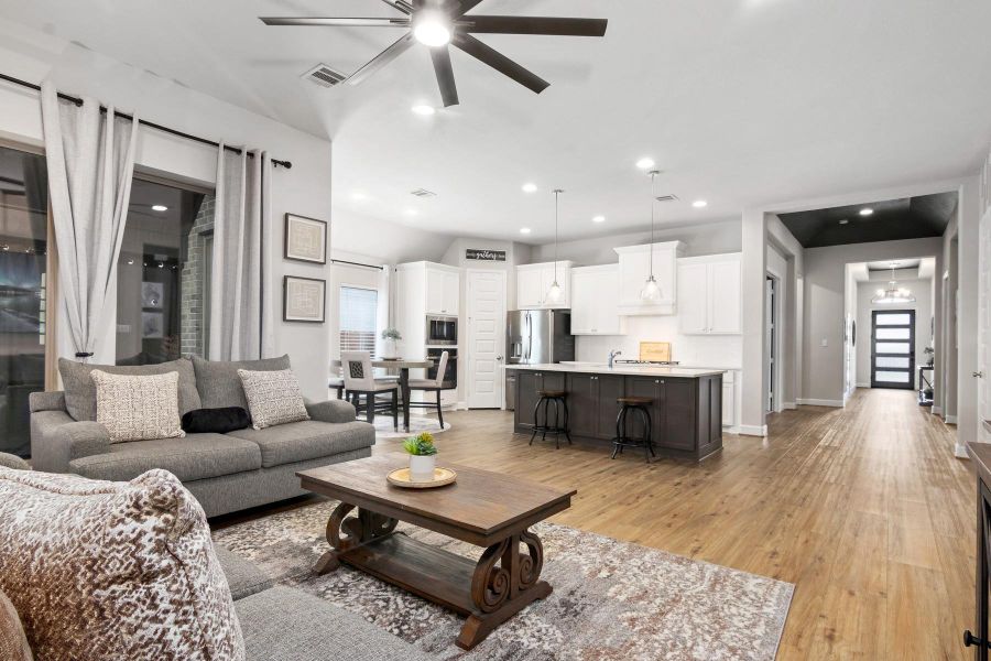 Spacious open-concept living area with modern kitchen, featuring white cabinetry, a central island with seating, and stainless steel appliances. The living room has a cozy seating arrangement, large windows with natural light, and elegant ceiling fan. Wood flooring extends throughout, creating a warm and inviting atmosphere.