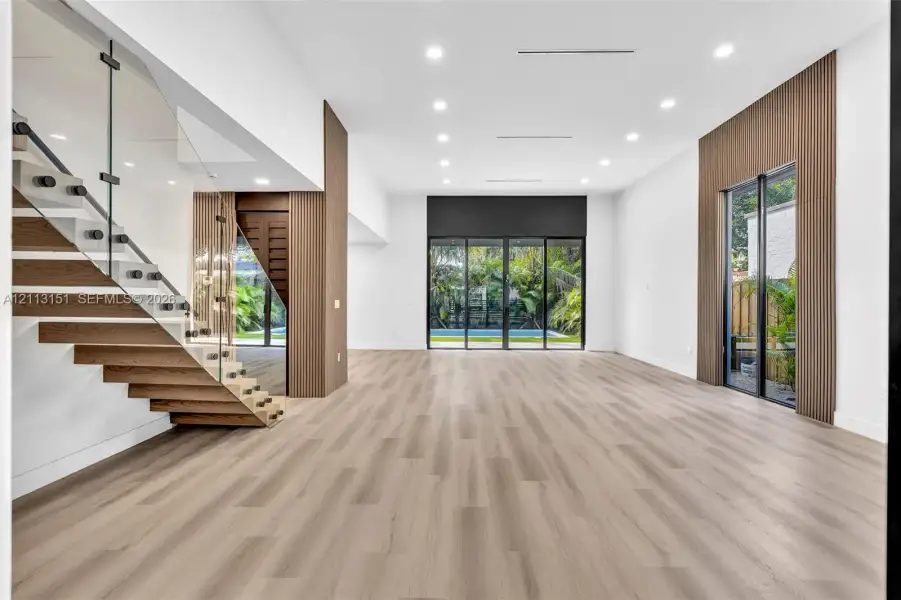 Open floor plan featuring wood-finish flooring, recessed lighting, floor-to-ceiling glass panel doors, and a glass-railed staircase with wood-finish treads and vertical wood wall slats Open floor plan featuring wood-finish flooring, recessed lighting, floor-to-ceiling glass panel doors, and a glass-railed staircase with wood-finish treads and vertical wood wall slats