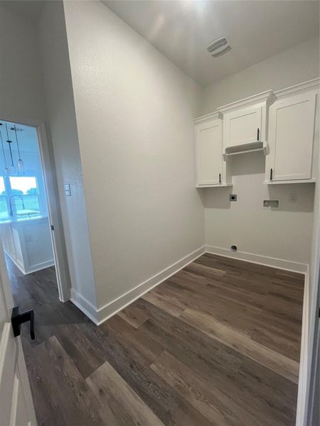 This photo shows a small laundry or utility room with light and medium gray/wood water proof plank flooring. It features white cabinetry and space for appliances, with an adjacent view into a well-lit kitchen area.