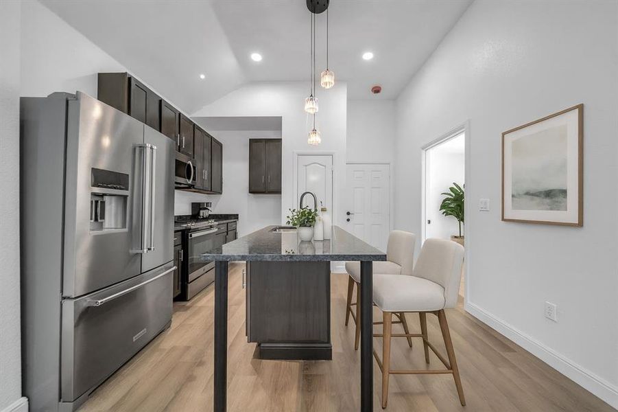 Staged - Kitchen featuring stainless steel appliances, dark brown cabinetry, pendant lighting, an island with sink, and dark stone counters