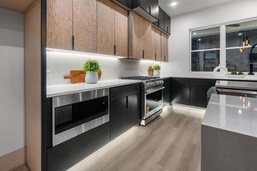 Kitchen with stainless steel appliances, dark cabinetry, light wood-style flooring, light stone counters, and modern cabinets