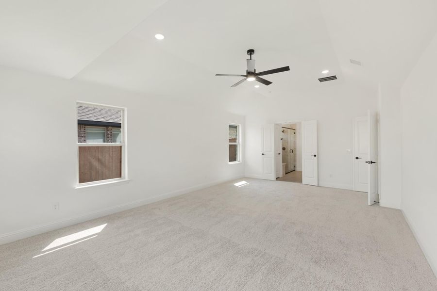 Spacious empty bedroom with vaulted ceiling, ceiling fan, carpet flooring, and open ensuite bathroom door