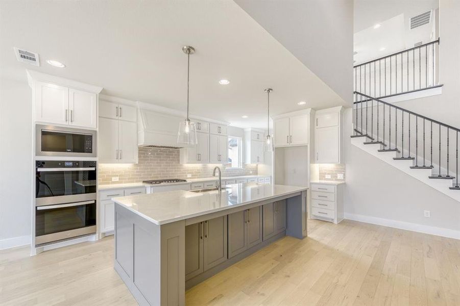 Kitchen with backsplash, light wood-type flooring, stainless steel appliances, pendant lighting, and a center island with sink