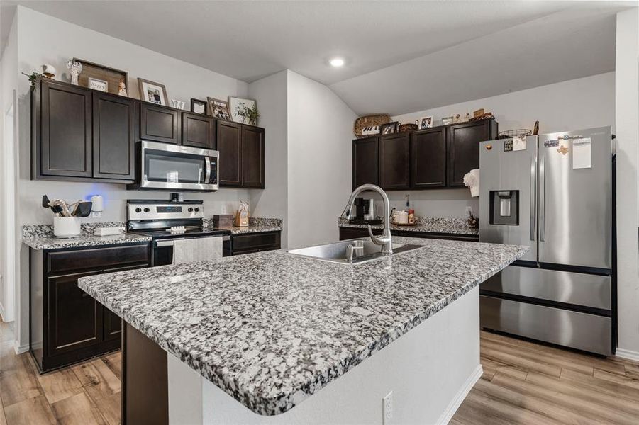 Kitchen with stainless steel appliances, light stone countertops, dark brown cabinets, light wood-style flooring, and a center island with sink