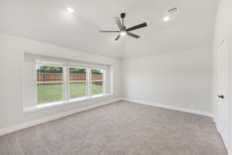 Empty bedroom with vaulted ceiling, ceiling fan, carpet flooring, and large window overlooking backyard