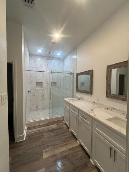 This bathroom features a spacious glass-enclosed shower with elegant tilework and dual shower niches. It includes a long, double-sink vanity with Quartz countertops and framed mirrors. The medium gray/wood water proof plank flooring adds warmth, and recessed lighting provides a bright, modern atmosphere.