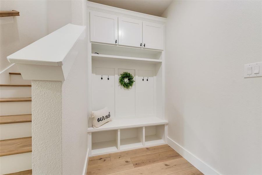 Built-in storage bench with upper cabinetry and coat hooks, featuring light wood-finish flooring and a white staircase with wood treads