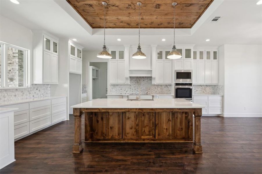 Kitchen with appliances with stainless steel finishes, a tray ceiling, dark wood-type flooring, hanging light fixtures, and an island with sink