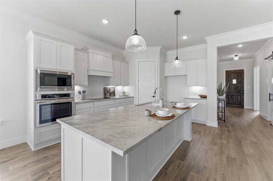 Contemporary kitchen featuring a large central island with a light-toned countertop, white cabinetry with crown molding, stainless steel built-in appliances, and wood-finish flooring