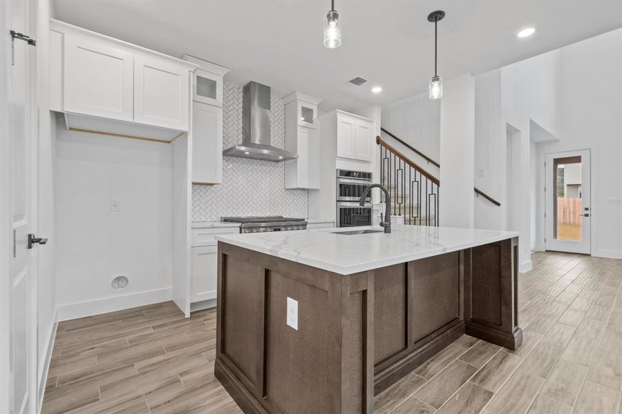 Kitchen with decorative light fixtures, white cabinets, wall chimney range hood, and a center island with sink