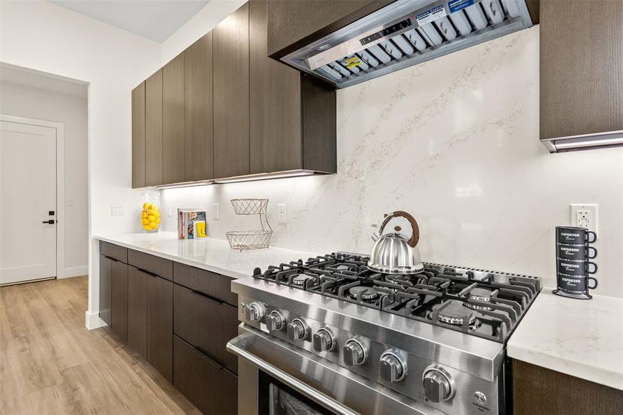 Gourmet kitchen featuring a stainless steel gas range, coordinating range hood, dark wood-finish cabinetry, light stone countertops, and a full-height stone slab backsplash