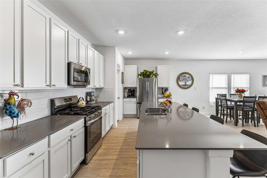 Modern kitchen featuring an expansive island with an undermount sink, white shaker-style cabinetry, stainless steel appliances, a subway tile backsplash, and wood-finish flooring Modern kitchen featuring an expansive island with an undermount sink, white shaker-style cabinetry, stainless steel appliances, a subway tile backsplash, and wood-finish flooring