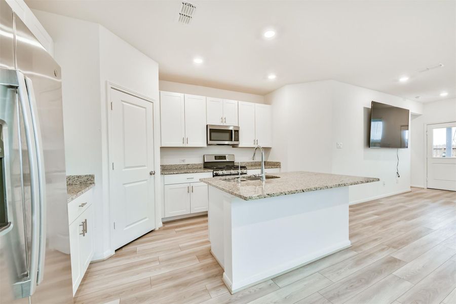 This modern kitchen features granite countertops, a central island with a sink, stainless steel appliances, and ample white cabinetry. It opens up to a living area with wood-style flooring.