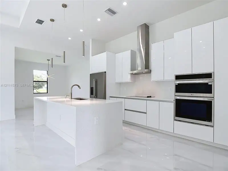 Modern kitchen featuring high-gloss cabinetry, stone-surface waterfall island with an undermount sink, built-in stainless steel double oven, stainless steel chimney range hood, and light-tone flooring