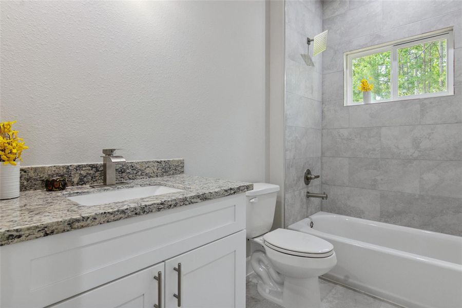 This bathroom features a modern design with a granite countertop, white cabinetry, and a sleek undermount sink. The shower-tub combo is surrounded by elegant gray tile, and a small window provides natural light.