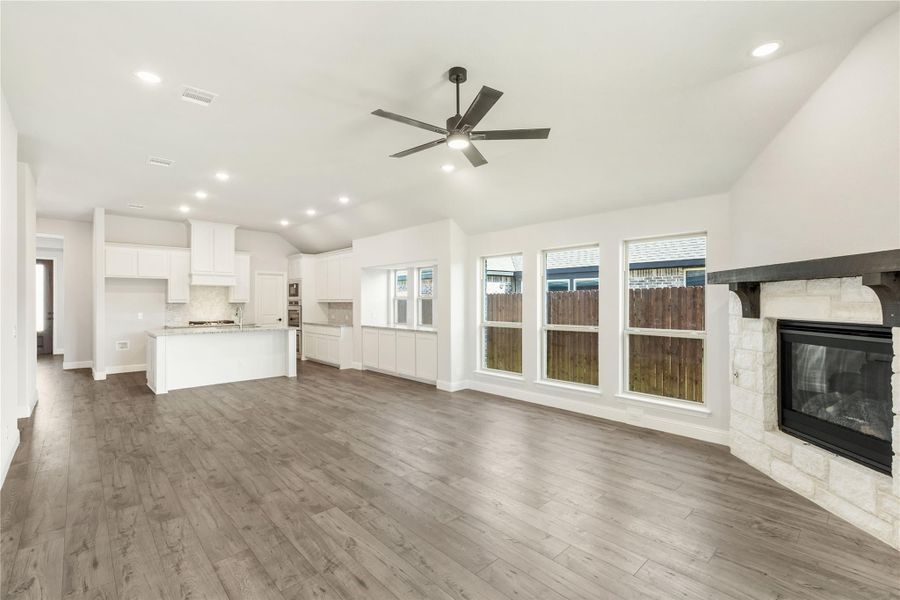 Open-concept living area with stone fireplace, hardwood floors, and white kitchen with island in background.