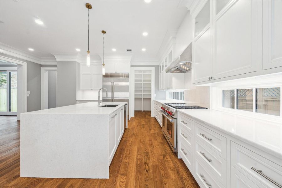 A closer look at the kitchen featuring sleek white cabinetry, a large island with sink and dishwasher, stainless steel appliances, including a Subzero refrigerator, Wolf gas stove, hardwood floors and a oversized walk-in pantry.  Natural light brightens the space through under counter windows, and sliding glass doors that open to the inviting outdoor space.