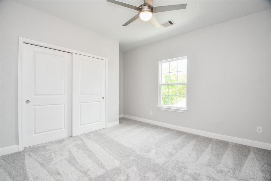 This is a bright, secondary room featuring neutral walls and carpeted flooring, with a window providing natural light and a ceiling fan for comfort. It includes a double-door closet for storage.