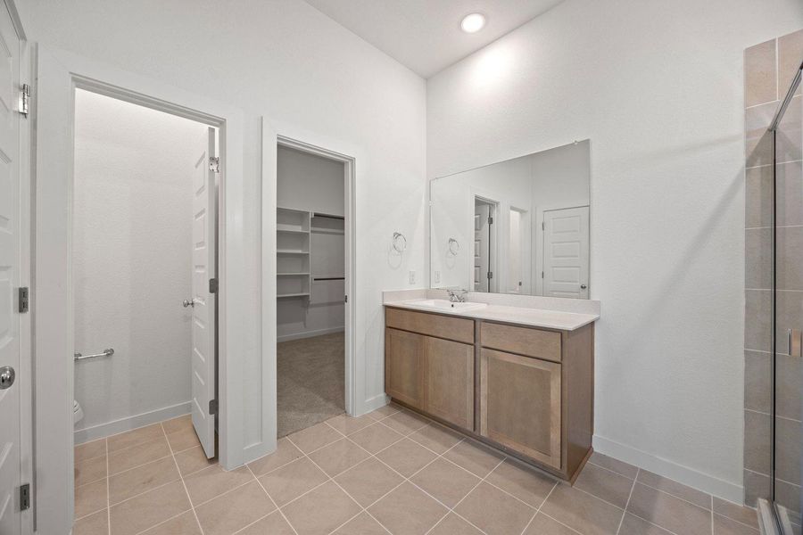 A well-lit primary bathroom featuring a single vanity, spacious mirror, and tiled shower.