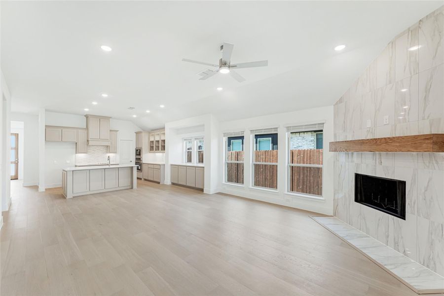 Open living area with marble tile fireplace, light wood floors, and kitchen with gray cabinets in background