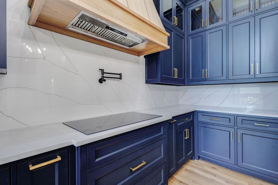 Kitchen featuring custom blue cabinetry with gold-tone hardware, a wood-finish range hood, white countertops, a contrasting white backsplash, and an integrated cooktop