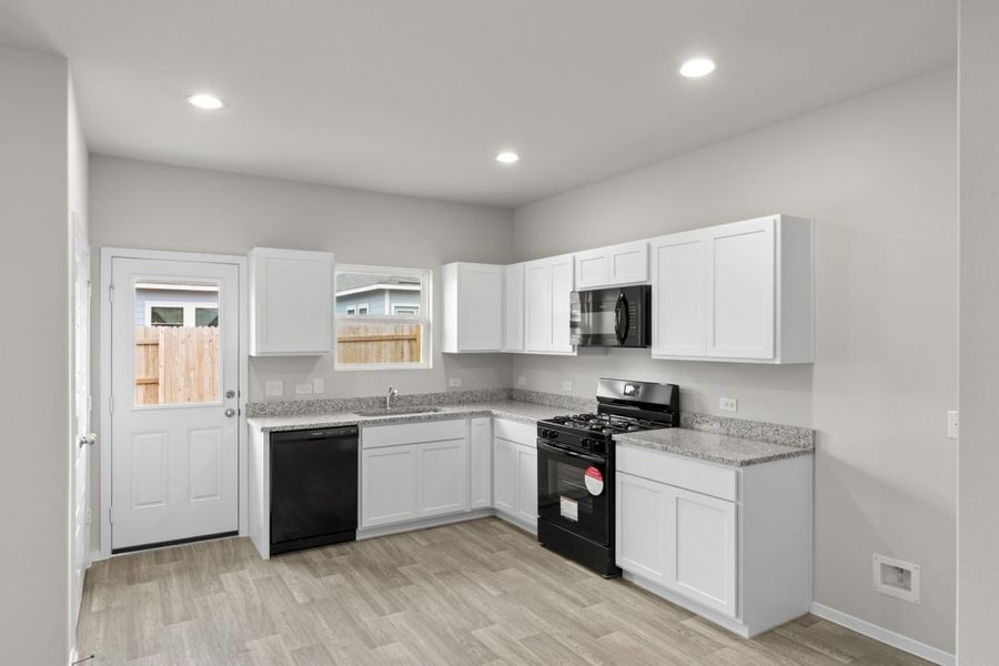 Image of an L-shaped kitchen with white cabinets, black appliances, a white back door and a window above the sink