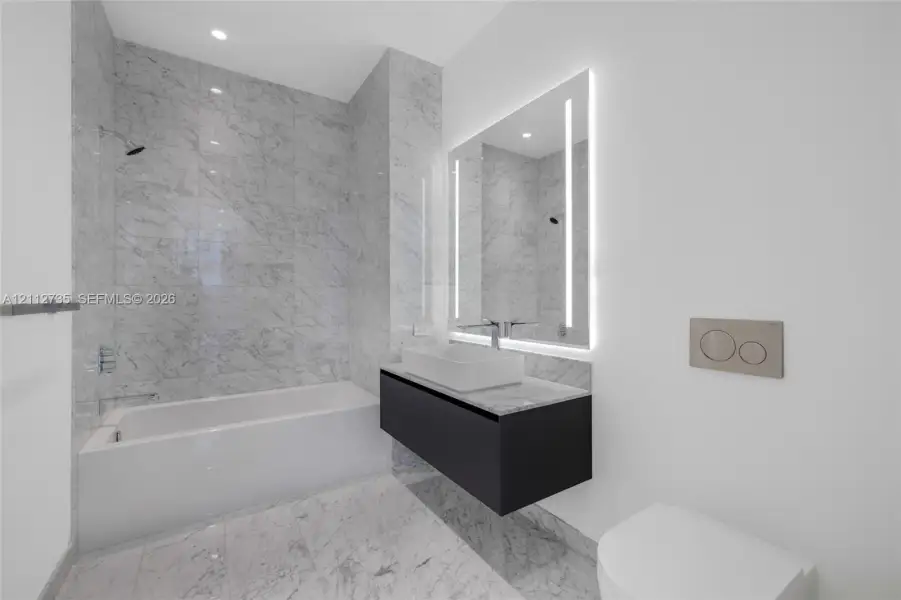 Full-height stone wall tiling, floating vanity with vessel sink, wall-mounted faucet, illuminated mirror, and wall-mounted dual-flush toilet