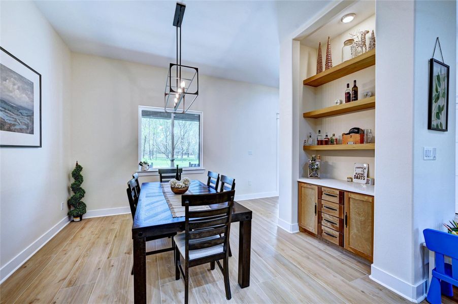 Bright dining area with light wood flooring and a modern rectangular table. A large window provides ample natural light, and an adjacent built-in shelving unit offers stylish storage. The room features a contemporary light fixture and neutral-colored walls.