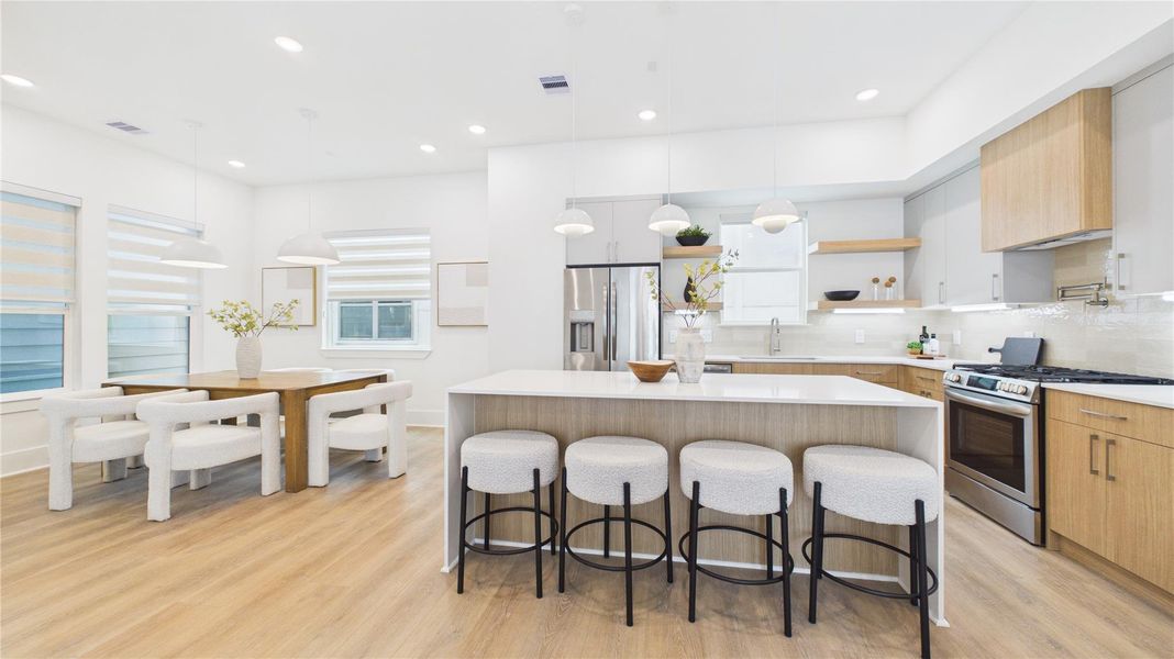 This photo showcases a modern, open-concept kitchen and dining area. The kitchen features a large island with seating, stainless steel appliances, and sleek cabinetry. The dining area has a stylish wooden table with cushioned chairs, and the space is bright with plenty of natural light and contemporary pendant lighting.