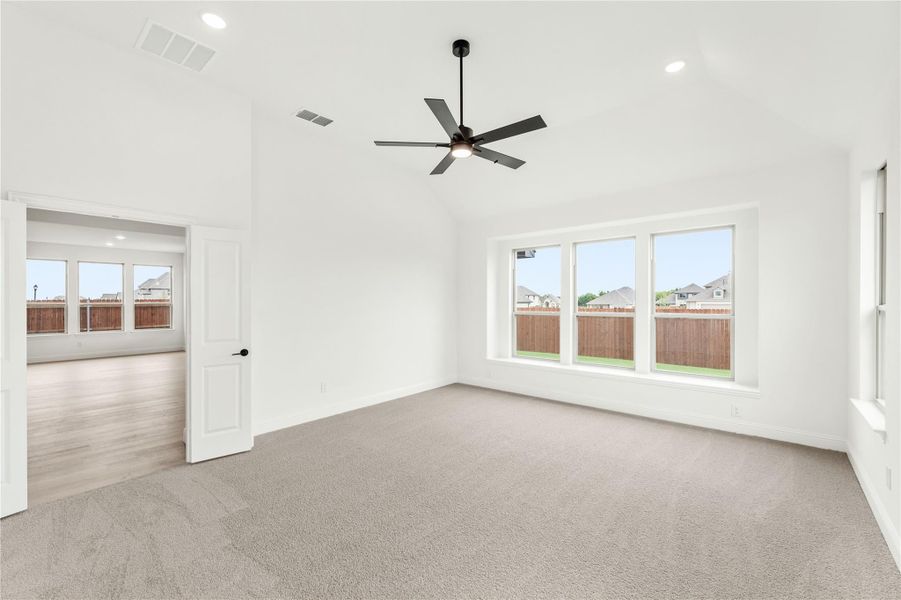 Spacious bedroom with vaulted ceiling, ceiling fan, carpet flooring, and large windows overlooking backyard fence Spacious bedroom with vaulted ceiling, ceiling fan, carpet flooring, and large windows overlooking backyard fence