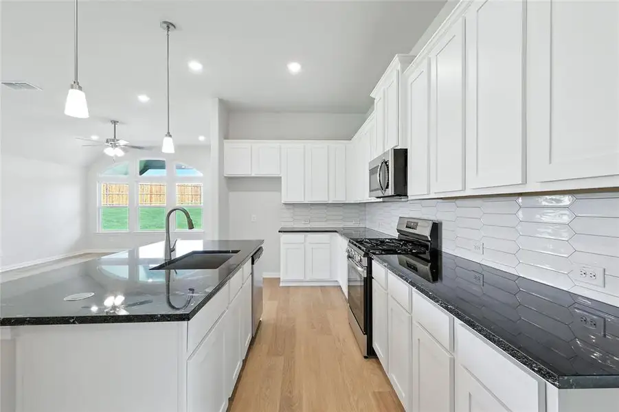 Kitchen featuring stainless steel appliances, dark stone countertops, a kitchen island with sink, light wood finished floors, and decorative light fixtures Kitchen featuring stainless steel appliances, dark stone countertops, a kitchen island with sink, light wood finished floors, and decorative light fixtures