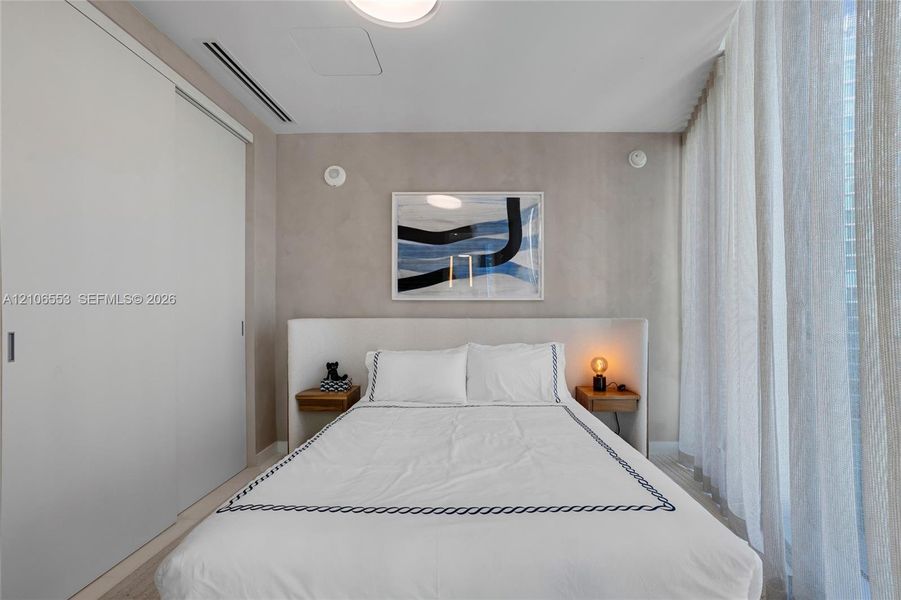 Neutral-toned room featuring a built-in upholstered headboard, dual integrated wood-finish bedside shelves, floor-to-ceiling sliding closet doors, and light-filtering floor-to-ceiling drapery