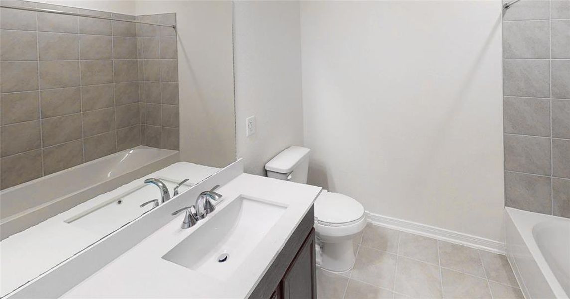 Secondary bath features tile flooring, bath/shower combo with tile surround, dark stained wood cabinets, beautiful light countertops, mirror, dark, sleek fixtures and modern finishes.