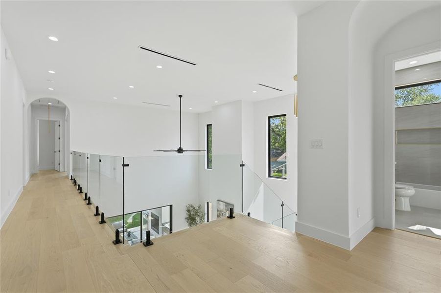 Open loft area featuring light wood-finish flooring, a glass railing with black posts, recessed ceiling lighting, a contemporary black ceiling fan, and an arched hallway entry Open loft area featuring light wood-finish flooring, a glass railing with black posts, recessed ceiling lighting, a contemporary black ceiling fan, and an arched hallway entry