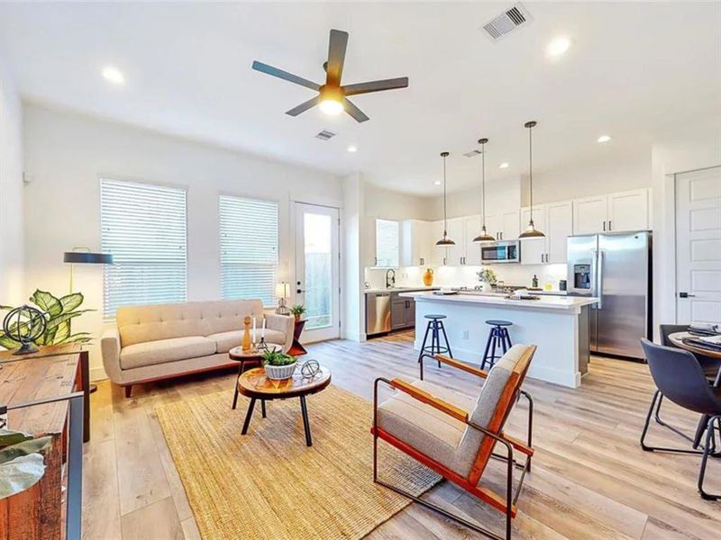 A modern and spacious living area with an open floor plan that seamlessly connects to the kitchen. Natural light floods the room, illuminating the warm wood floors and the contemporary furniture.