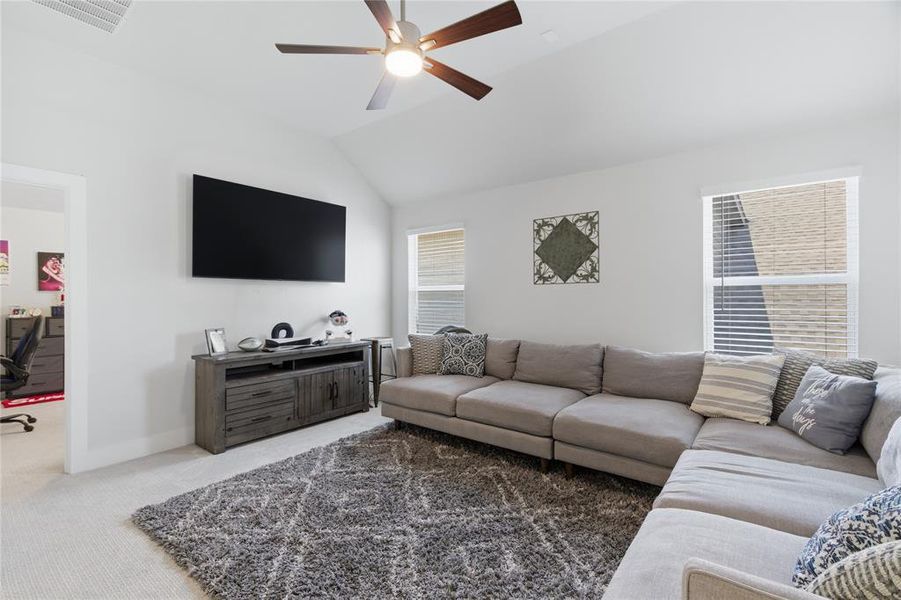 Spacious upstairs bonus room featuring a vaulted ceiling with a ceiling fan, two windows with blinds, a wall-mounted television, and light-colored carpeting