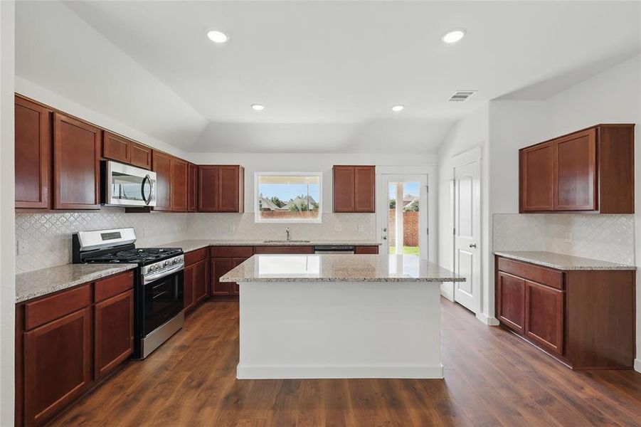 Kitchen with vaulted ceiling, stainless steel appliances, light stone counters, backsplash, and a center island