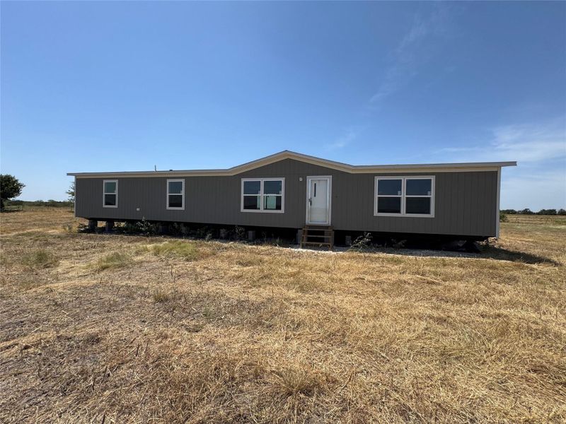A manufactured home stands on a sprawling rural lot under clear blue skies.