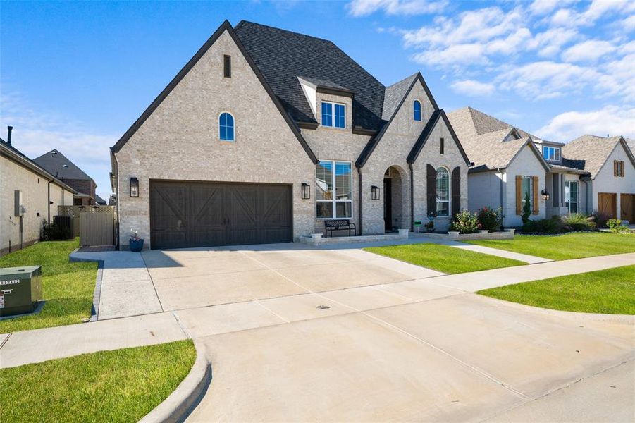 French country inspired facade featuring brick siding, concrete driveway, a garage, and roof with shingles French country inspired facade featuring brick siding, concrete driveway, a garage, and roof with shingles