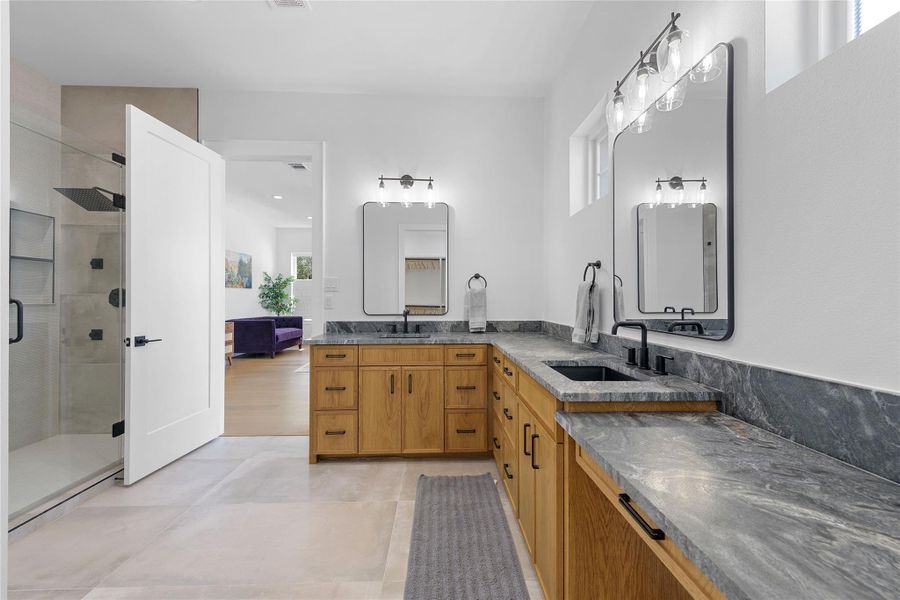 Designer finishes elevate this bathroom with custom wood cabinetry, dramatic natural stone countertops, dual vanities, matte black fixtures, and an oversized walk-in shower with sleek glass enclosure. Designer finishes elevate this bathroom with custom wood cabinetry, dramatic natural stone countertops, dual vanities, matte black fixtures, and an oversized walk-in shower with sleek glass enclosure.
