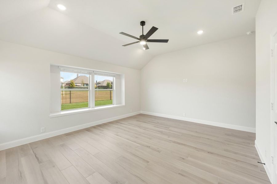 Empty bedroom with vaulted ceiling, ceiling fan, light wood floors, and large window overlooking backyard