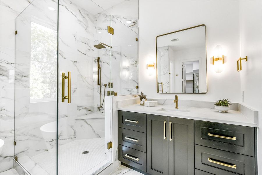 From this refined angle, the focus falls on the precision tile layout and architectural detailing. The frameless glass enclosure gleams against the veined marble tile, housing rainfall and handheld fixtures in brushed brass. The adjacent vanity continues the theme of bespoke craftsmanship, framed by sconce lighting and a minimalist aesthetic that balances opulence with calmsimplicity.
