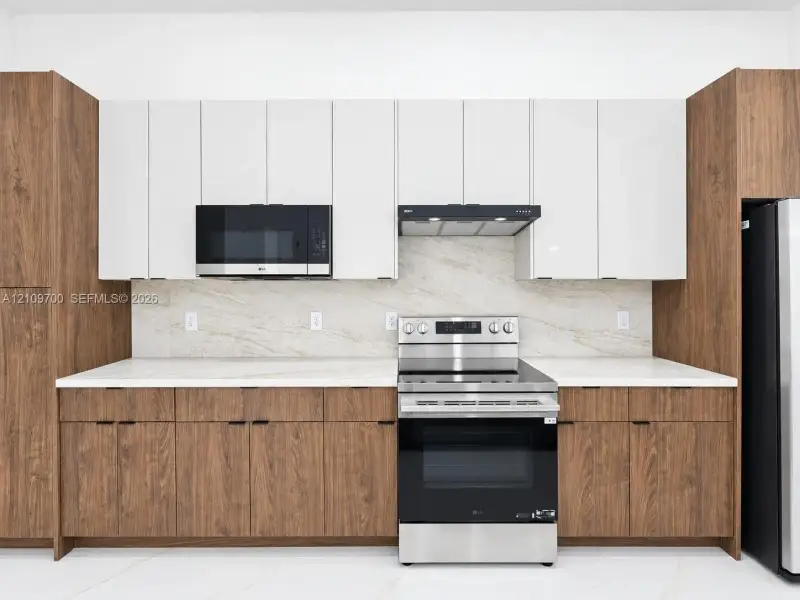 Contemporary kitchen featuring dual-tone cabinetry, stone-finish backsplash, white stone-composite countertops, and stainless steel appliances