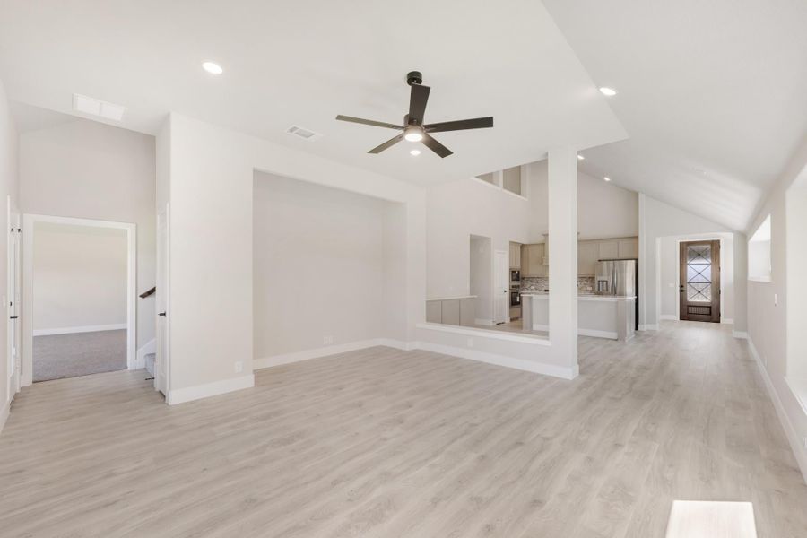 Open living area with vaulted ceiling, ceiling fan, light wood floors, and open view to kitchen beyond.