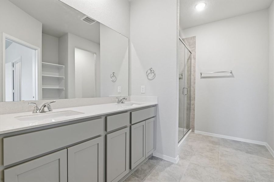 Image of a primary bathroom with light grey cabinets, a white jack and jill vanity, a large mirror and a walk-in shower